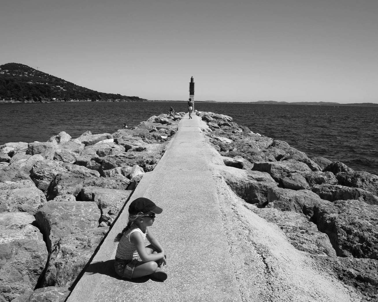 petite fille assise sur la jetée et qui regarde la mer phare au loin mer calme photo noir et blanc bnw
